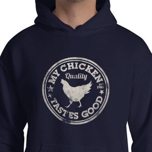 Könnte beinhalten: Ein marineblauer Hoodie mit einem weißen, abgenutzten Kreis-Motiv, auf dem "My Chicken Quality Est. 1985 Tastes Good" steht und sich eine Hühnersilhouette im Kreis befindet.