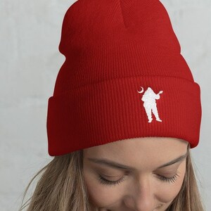 Peut inclure: Un bonnet rouge avec un motif brodé blanc représentant une silhouette jouant de la guitare sous un croissant de lune. Le bonnet est en tricot doux et possède un revers. Un accessoire d'hiver.