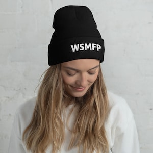 May include: Black beanie with the white letters "WSMFP" embroidered on the front. The beanie is made of a soft material and is worn by a person with long blonde hair. The background is a white brick wall.