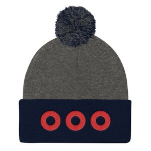 Puede incluir: Un gorro gris con un borde azul marino. El borde presenta tres círculos rojos bordados. Un pompón gris oscuro se encuentra en la parte superior del gorro. Este accesorio de invierno es elegante y adecuado para todos.