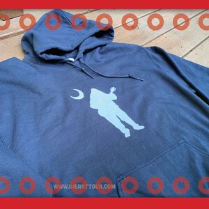 May include: A dark blue hooded sweatshirt with a white silhouette of a person playing a guitar and a crescent moon. The text "www.jhearttour.com" is printed on the bottom of the sweatshirt.