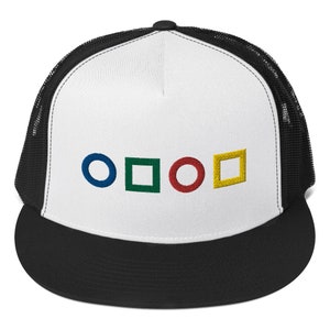 Puede incluir: Una gorra de camionero negra y blanca con un panel frontal blanco que presenta cuatro símbolos bordados: un círculo azul, un cuadrado verde, un círculo rojo y un cuadrado amarillo.