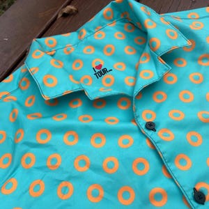May include: A turquoise blue button-up shirt with a pattern of orange donut rings. The shirt has a small embroidered patch on the left side of the chest that says "I LOVE TOUR..."