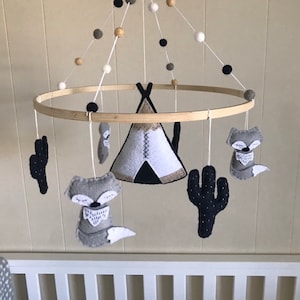 K&ouml;nnte beinhalten: Ein Holz-Babymobil mit einem Tipi, zwei F&uuml;chsen und zwei Kakteen, die an einem runden Holzrahmen h&auml;ngen. Das Mobile ist mit wei&szlig;en, schwarzen und beigefarbenen Pompons verziert.