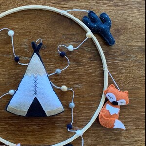 K&ouml;nnte beinhalten: Ein Mobile aus Holz mit Filztieren und geometrischen Formen. Das Mobile zeigt ein blaues und wei&szlig;es Tipi, einen schlafenden orangefarbenen Fuchs und einen dunkelblauen Kaktus. Das Mobile ist mit wei&szlig;em Garn und wei&szlig;en Pompons bespannt.