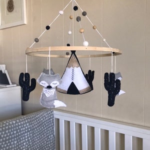 K&ouml;nnte beinhalten: Ein Holz-Babymobil mit einem Tipi, zwei F&uuml;chsen und zwei Kakteen, die daran h&auml;ngen. Das Mobile ist mit wei&szlig;en, schwarzen und beigefarbenen Filzkugeln verziert.