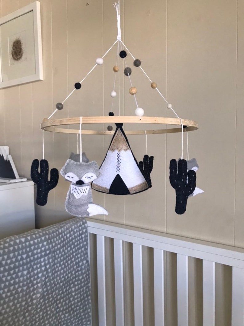 K&ouml;nnte beinhalten: Ein Holz-Babymobil mit einem Tipi, zwei F&uuml;chsen und zwei Kakteen, die daran h&auml;ngen. Das Mobile ist mit wei&szlig;en, schwarzen und beigefarbenen Filzkugeln verziert.