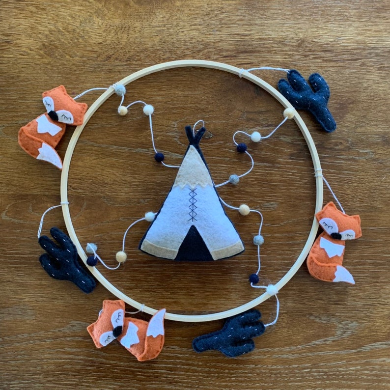 K&ouml;nnte beinhalten: Ein Mobile aus Holz mit Filztier- und Pflanzendekorationen. Das Mobile zeigt ein Tipi, zwei F&uuml;chse und zwei Kakteen. Das Tipi ist wei&szlig; mit schwarzem Besatz und hat eine braune Filzbasis. Die F&uuml;chse sind orange mit schwarzen Augen und Nasen. Die Kakteen sind gr&uuml;n mit schwarzen Stacheln.