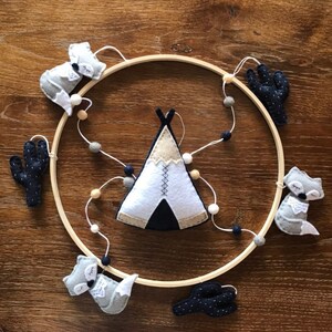 K&ouml;nnte beinhalten: Ein Mobile aus Holz mit einem Kreis als Rahmen und Filzfiguren von Tieren und Kakteen. Das Mobile zeigt ein wei&szlig;es Tipi mit schwarzen und braunen Akzenten, drei graue F&uuml;chse und zwei schwarze Kakteen mit wei&szlig;en Punkten.