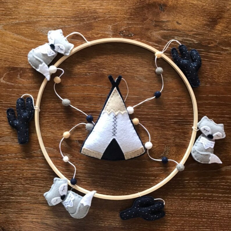 K&ouml;nnte beinhalten: Ein Mobile aus Holz mit einem Kreis als Rahmen und Filzfiguren von Tieren und Kakteen. Das Mobile zeigt ein wei&szlig;es Tipi mit schwarzen und braunen Akzenten, drei graue F&uuml;chse und zwei schwarze Kakteen mit wei&szlig;en Punkten.