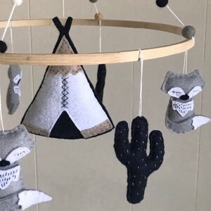 K&ouml;nnte beinhalten: Ein Baby-Mobile mit einem Tipi, zwei F&uuml;chsen, einem Kaktus und Holzperlen. Das Tipi ist wei&szlig; mit schwarzen und braunen Akzenten. Die F&uuml;chse sind grau mit schwarzen Akzenten. Der Kaktus ist schwarz mit wei&szlig;en Akzenten. Die Holzperlen sind braun.