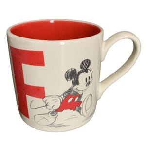 Puede incluir: Taza de cerámica color crema con interior rojo y una gran letra "F" roja en el lateral. La taza presenta un boceto de Mickey Mouse en negro, rojo y blanco. La taza tiene un asa curvada.