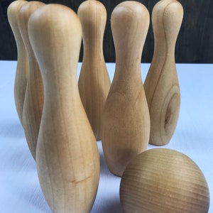 May include: A set of wooden bowling pins and a wooden bowling ball. The pins are arranged in a triangle formation, with the ball in front of them. The pins and ball are all a light brown color.