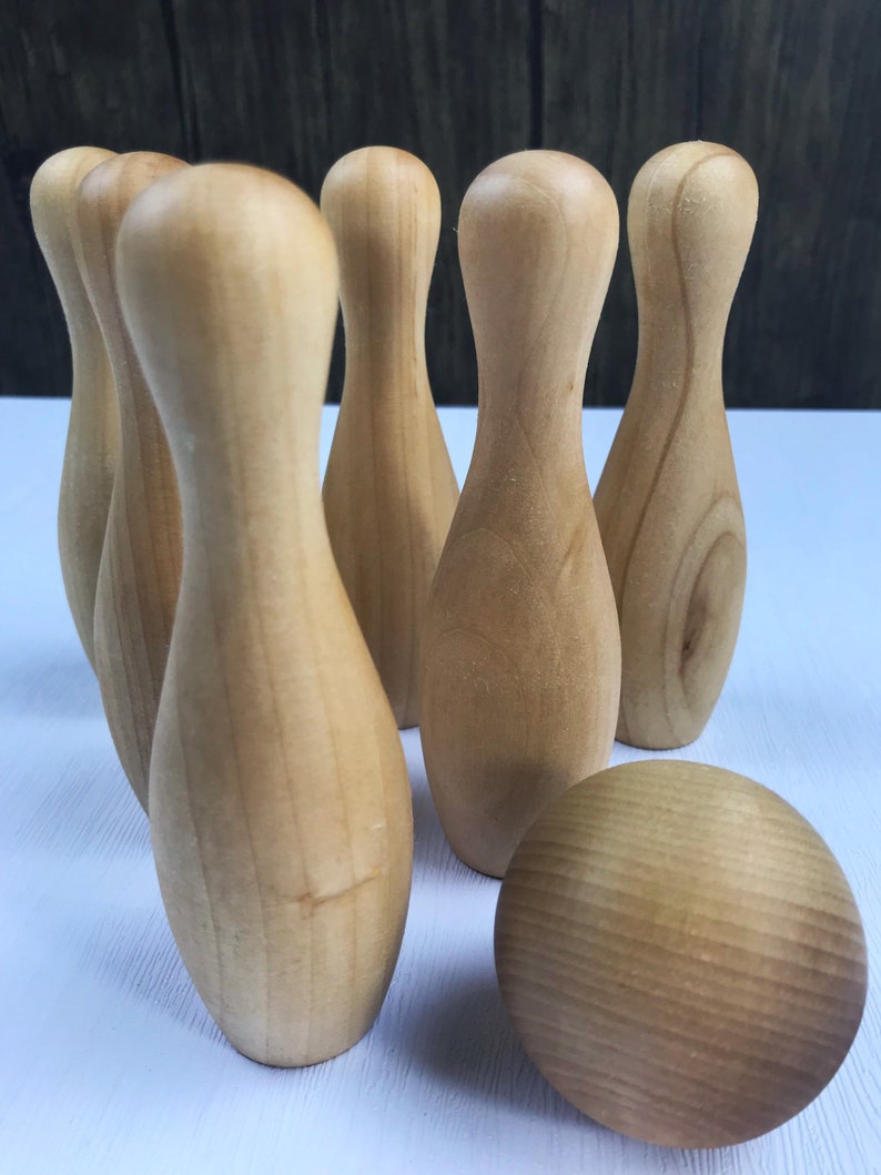 May include: A set of wooden bowling pins and a wooden bowling ball. The pins are arranged in a triangle formation, with the ball in front of them. The pins and ball are all a light brown color.