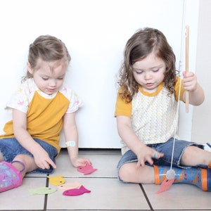 May include: Two children play a fishing game with felt fish on a tiled floor. One child holds a fishing rod with a magnet on the end, while the other child places the felt fish on the floor. The fish are in various colors, including pink, yellow, green, and orange.