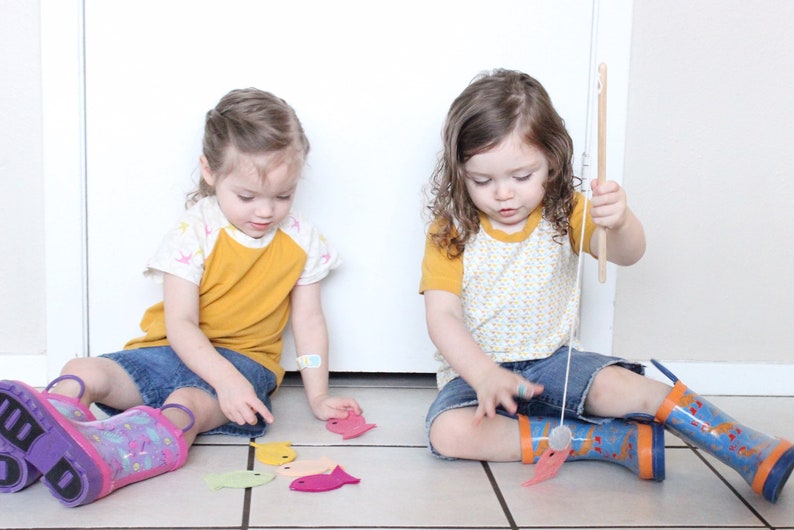 May include: Two children play a fishing game with felt fish on a tiled floor. One child holds a fishing rod with a magnet on the end, while the other child places the felt fish on the floor. The fish are in various colors, including pink, yellow, green, and orange.