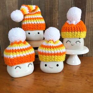 Puede incluir: Cuatro tazas de cer&aacute;mica blanca con caras lindas, cada una con un gorro de punto a rayas naranja, amarillo y blanco hecho a mano con un pomp&oacute;n blanco.