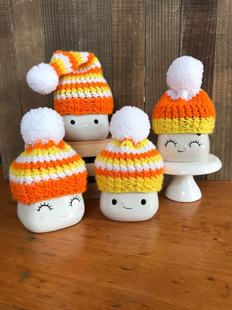 Puede incluir: Cuatro tazas de cer&aacute;mica blanca con caras lindas, cada una con un gorro de punto a rayas naranja, amarillo y blanco hecho a mano con un pomp&oacute;n blanco.