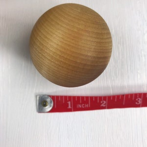 May include: A close-up of a light brown wooden ball, approximately 1 inch in diameter, with a smooth, slightly textured surface. The ball is sitting on a white surface.