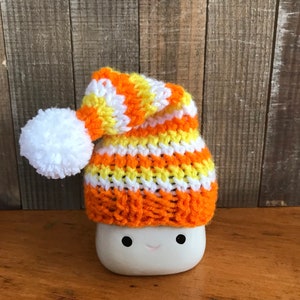 Puede incluir: Una taza de cer&aacute;mica blanca con una cara sonriente que lleva un gorro de punto con rayas naranjas, amarillas y blancas. El gorro tiene un pomp&oacute;n blanco en la parte superior.