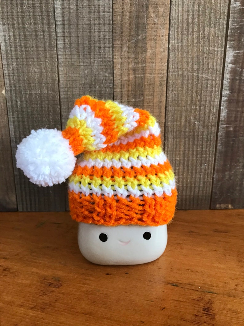 Puede incluir: Una taza de cer&aacute;mica blanca con una cara sonriente que lleva un gorro de punto con rayas naranjas, amarillas y blancas. El gorro tiene un pomp&oacute;n blanco en la parte superior.