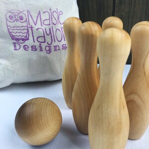 May include: A set of wooden bowling pins and a ball, all in a natural wood color. The pins are arranged in a triangle shape, with the ball in front of them. The set is on a white surface.