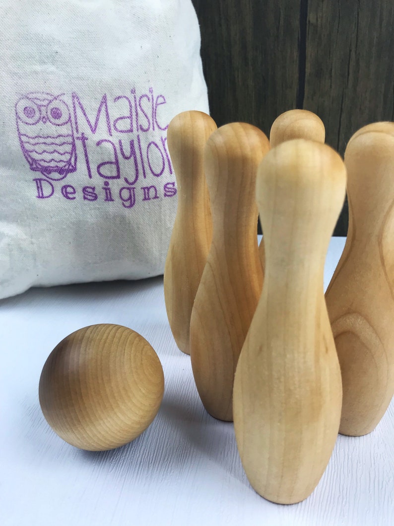 May include: A set of wooden bowling pins and a ball, all in a natural wood color. The pins are arranged in a triangle shape, with the ball in front of them. The set is on a white surface.