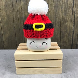 May include: A small, white ceramic mug with a smiling face, topped with a handmade red and black Santa hat with a yellow buckle and a white pom-pom. The mug sits on a small wooden crate, perfect for holiday decor.