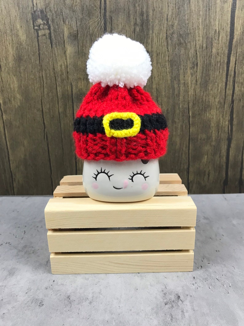 May include: A small, white ceramic mug with a smiling face, topped with a handmade red and black Santa hat with a yellow buckle and a white pom-pom. The mug sits on a small wooden crate, perfect for holiday decor.