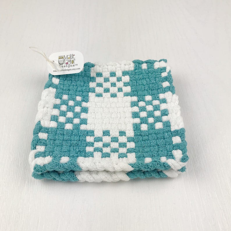May include: A folded, square, woven cloth in teal and white. The pattern is a repeating checkerboard design. A small tag with an owl logo and text is attached to the cloth.