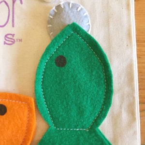 May include: A green felt fish with a black eye, stitched with white thread, is shown in close-up. Above the fish is a grey felt circle, attached with a white string. The fish is on cream-coloured fabric.