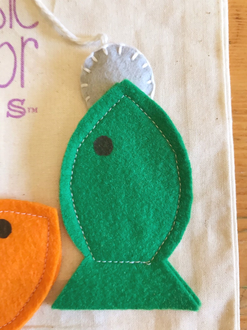 May include: A green felt fish with a black eye, stitched with white thread, is shown in close-up. Above the fish is a grey felt circle, attached with a white string. The fish is on cream-coloured fabric.