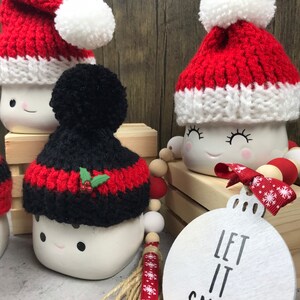 May include: White ceramic mugs with smiling faces, each adorned with a knitted Santa hat in red and white or black and red. A wooden ornament with "LET IT SNOW" text and a beaded garland complete the festive scene.