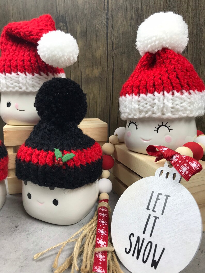 May include: White ceramic mugs with smiling faces, each adorned with a knitted Santa hat in red and white or black and red. A wooden ornament with "LET IT SNOW" text and a beaded garland complete the festive scene.