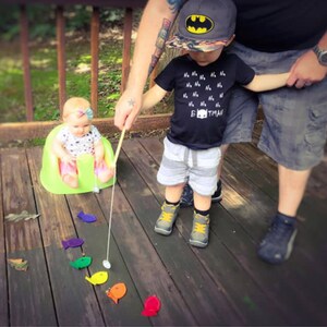 May include: A child in a Batman cap and a black t-shirt with the text "Batman" plays with a fishing rod. A baby sits in a green chair. Brightly coloured fish-shaped toys are scattered on a wooden deck.