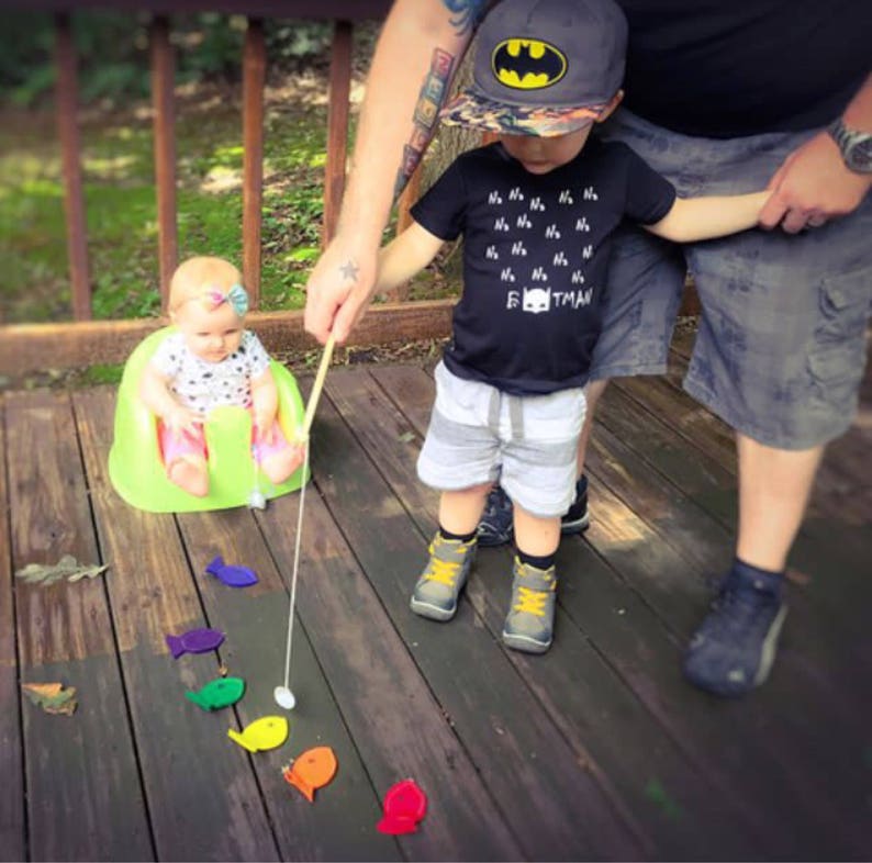 May include: A child in a Batman cap and a black t-shirt with the text "Batman" plays with a fishing rod. A baby sits in a green chair. Brightly coloured fish-shaped toys are scattered on a wooden deck.