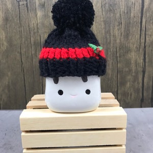 May include: A white ceramic marshmallow figure with black eyes and a smiling mouth, wearing a black and red knit hat with a pom-pom and a small holly accent. The figure is displayed on a small wooden crate.