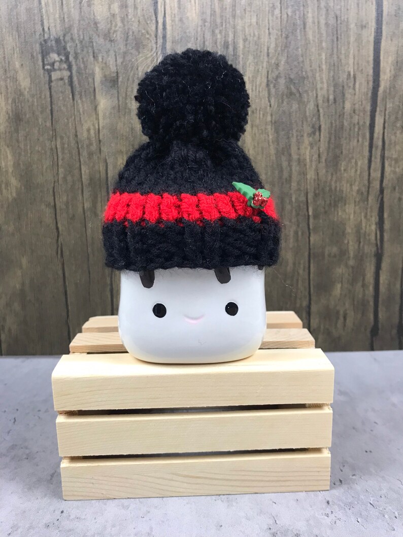 May include: A white ceramic marshmallow figure with black eyes and a smiling mouth, wearing a black and red knit hat with a pom-pom and a small holly accent. The figure is displayed on a small wooden crate.