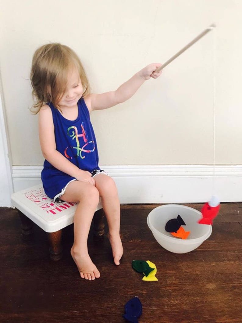 May include: A child in a blue dress plays with a fishing rod and felt fish. The child sits on a small white stool. A white bowl contains felt fish in various colours. The floor is made of dark wood.