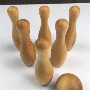 May include: A set of ten wooden bowling pins and a wooden bowling ball. The pins are arranged in a triangle formation, with the ball in front of them. The pins and ball are all a light brown color.