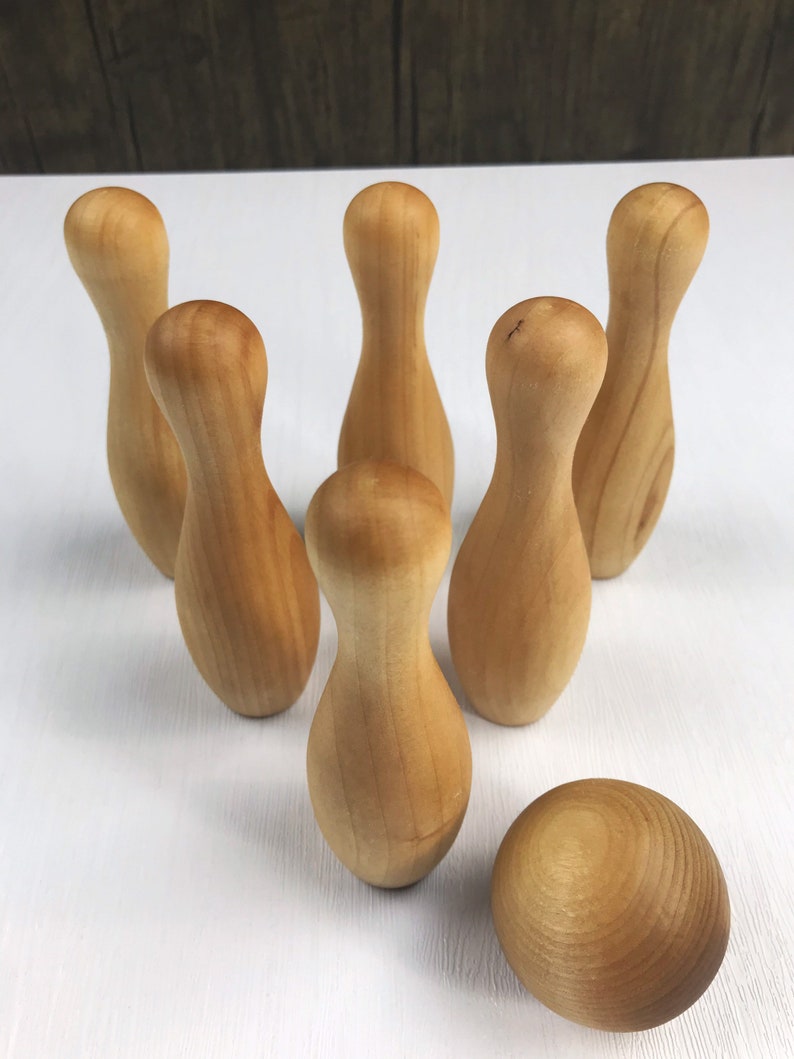 May include: A set of ten wooden bowling pins and a wooden bowling ball. The pins are arranged in a triangle formation, with the ball in front of them. The pins and ball are all a light brown color.