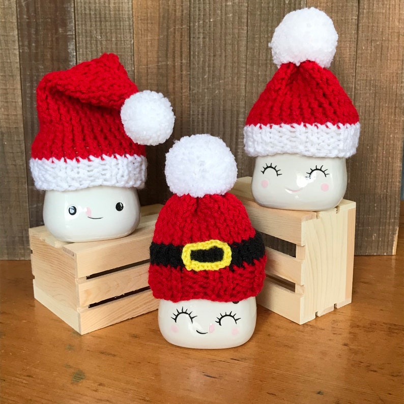 May include: Three white ceramic mugs with smiling faces, each adorned with a crocheted Santa hat. The hats are primarily red with white trim and a white pom-pom. One hat features a black belt with a yellow buckle. The mugs are displayed on a wooden crate.