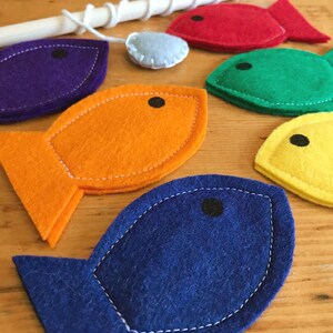 May include: A children's fishing game featuring felt fish in vibrant colours: purple, orange, red, green, yellow, and blue. The game includes a wooden rod and a small, light blue felt piece. Each fish has a black eye and white stitching.