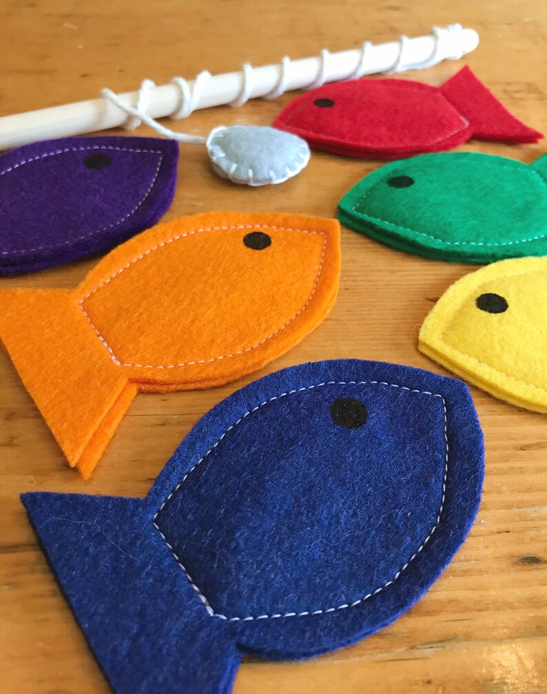 May include: A children's fishing game featuring felt fish in vibrant colours: purple, orange, red, green, yellow, and blue. The game includes a wooden rod and a small, light blue felt piece. Each fish has a black eye and white stitching.
