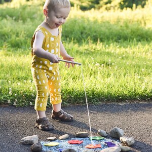 May include: A child in a yellow jumpsuit with a white moon pattern plays with a fishing rod and colourful fish cutouts arranged in a circle of stones on a paved surface. The child wears brown sandals.
