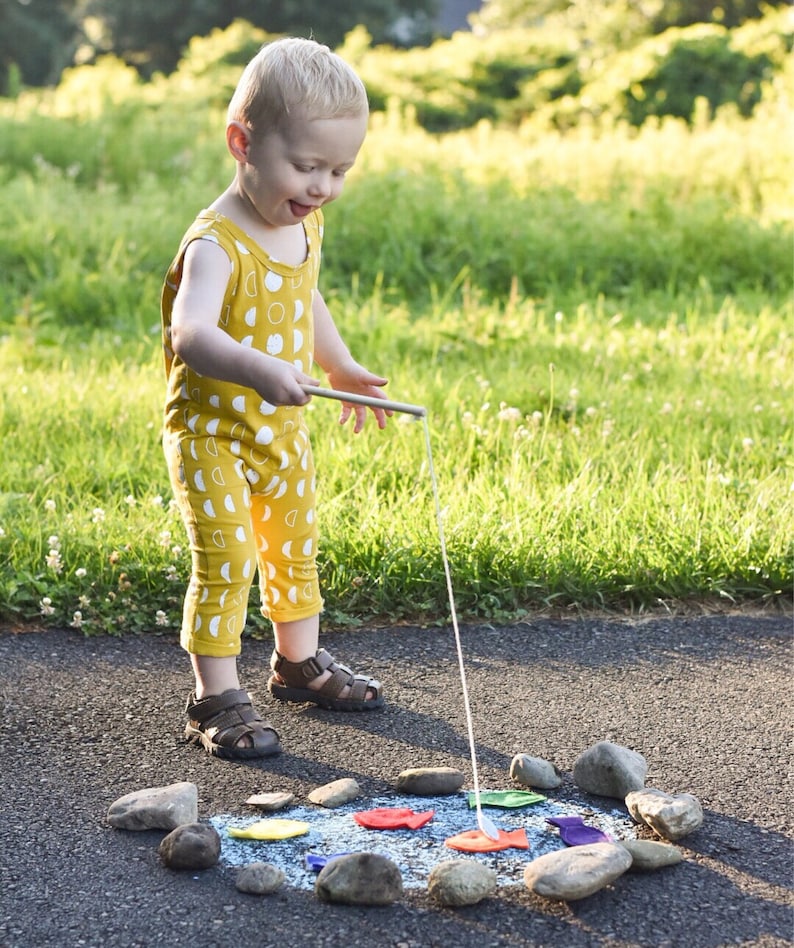 May include: A child in a yellow jumpsuit with a white moon pattern plays with a fishing rod and colourful fish cutouts arranged in a circle of stones on a paved surface. The child wears brown sandals.