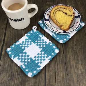 May include: A white mug with the word "FRIEND" printed on it, a slice of cake on a plate, and two blue and white checkered crochet potholders. The potholders are square and have a loop for hanging.