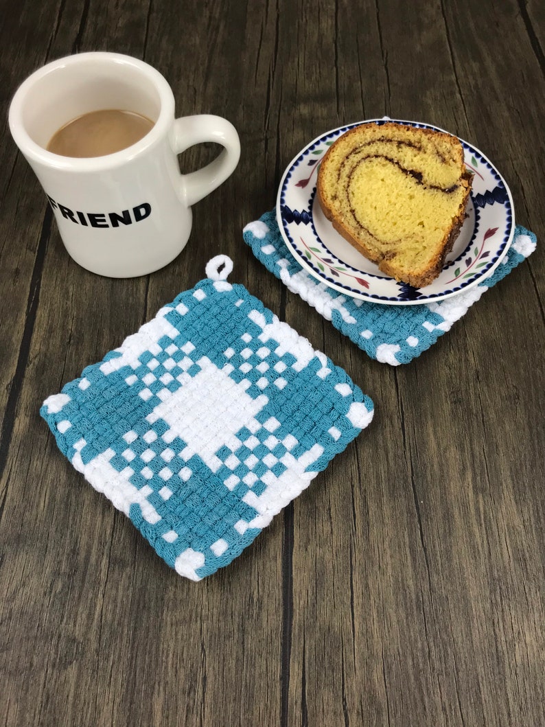 May include: A white mug with the word "FRIEND" printed on it, a slice of cake on a plate, and two blue and white checkered crochet potholders. The potholders are square and have a loop for hanging.