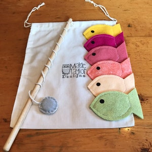 May include: A felt fishing game with a wooden rod, a gray felt circle, and five felt fish in different colors: yellow, pink, red, orange, and green. The fish are attached to the rod with string. The game is packaged in a white drawstring bag with the text "Maisie Taylor Designs" and a small owl logo.