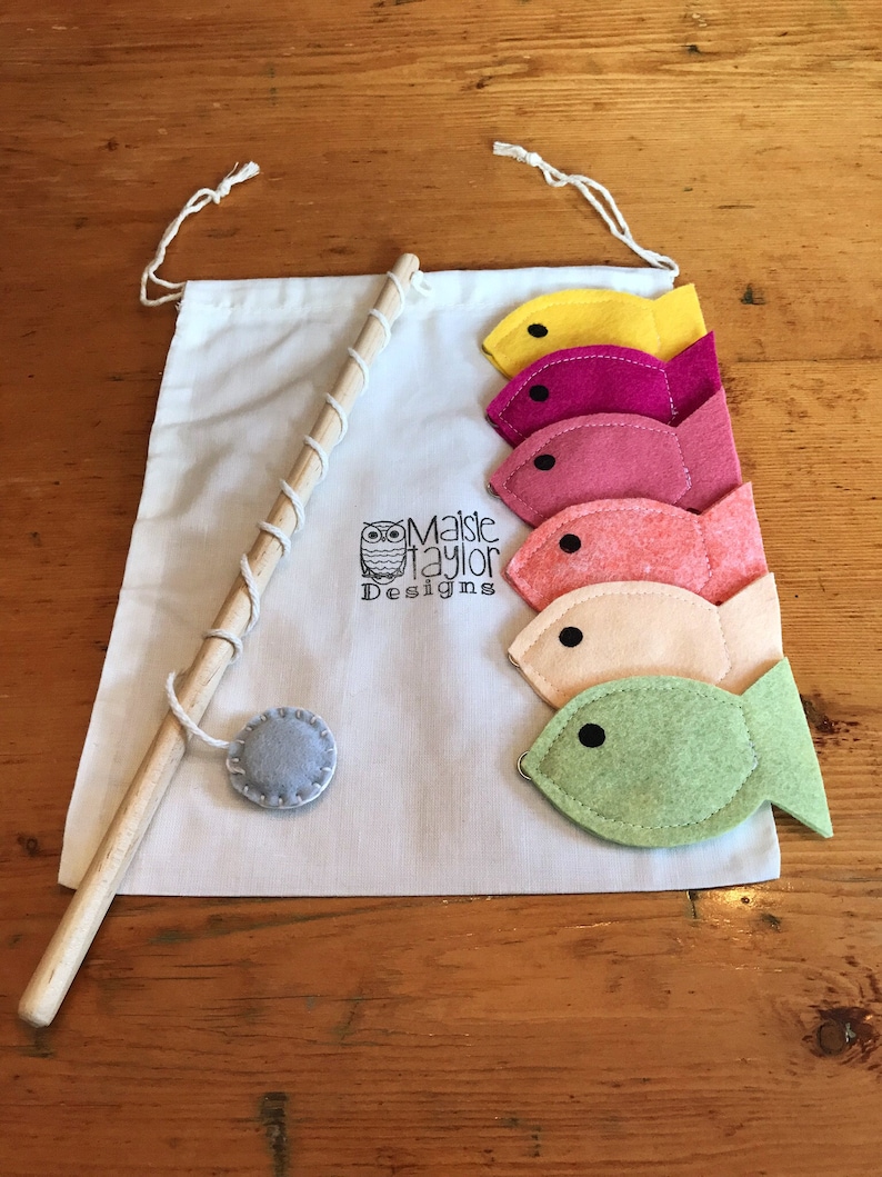 May include: A felt fishing game with a wooden rod, a gray felt circle, and five felt fish in different colors: yellow, pink, red, orange, and green. The fish are attached to the rod with string. The game is packaged in a white drawstring bag with the text "Maisie Taylor Designs" and a small owl logo.